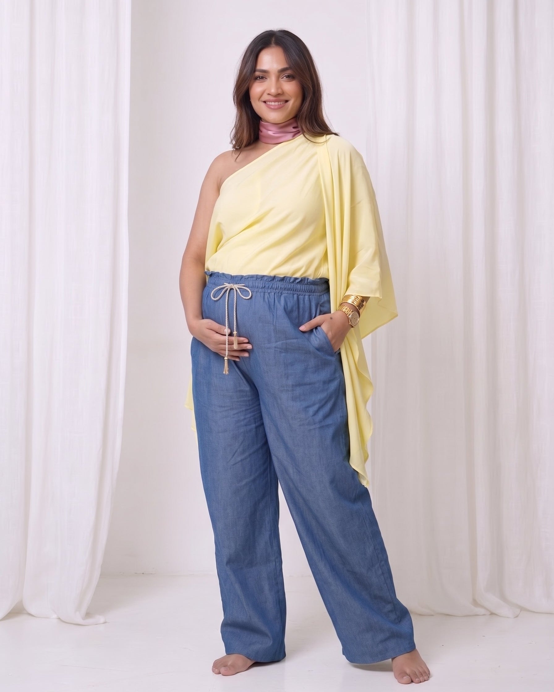 A woman modeling a two-piece outfit consisting of a pale yellow draped one-shoulder top and wide-leg blue drawstring pants, standing against a white draped fabric backdrop.