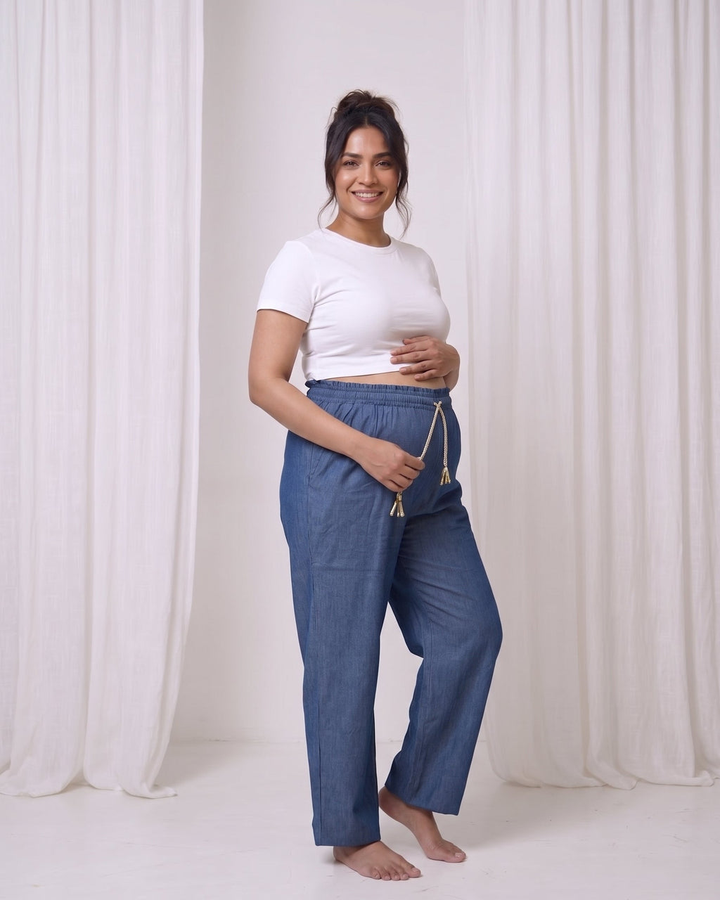 A pregnant woman stands barefoot in a studio wearing blue maternity lounge pants with a drawstring and a white cropped t-shirt, set against white draped curtains.