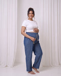 A pregnant woman stands barefoot in a studio wearing blue maternity lounge pants with a drawstring and a white cropped t-shirt, set against white draped curtains.