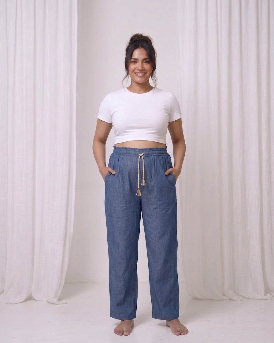 A woman stands barefoot in a studio wearing loose blue drawstring pants with a white cropped t-shirt, posing in front of white draped curtains.