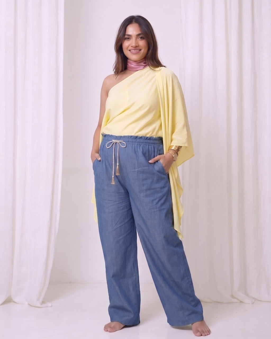 A woman modeling a two-piece outfit consisting of a pale yellow one-shoulder draped top and wide-leg blue pants, standing against a white draped fabric backdrop.