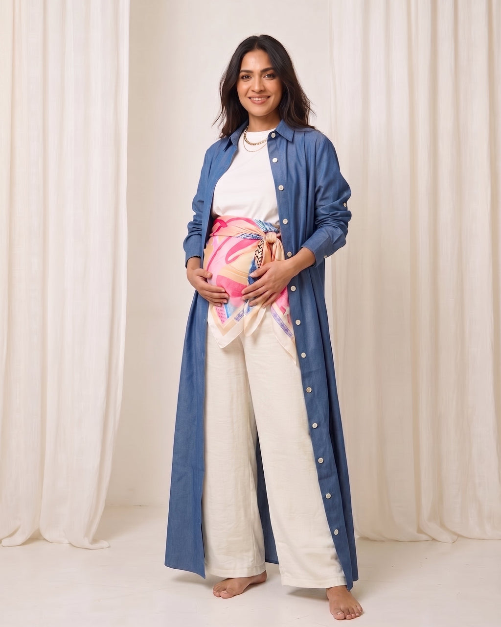 A pregnant woman stands barefoot in front of a cream-colored curtain backdrop, wearing a long blue button-up shirt over a white top and cream wide-leg pants, with a pink and blue patterned scarf wrapped around her belly.
