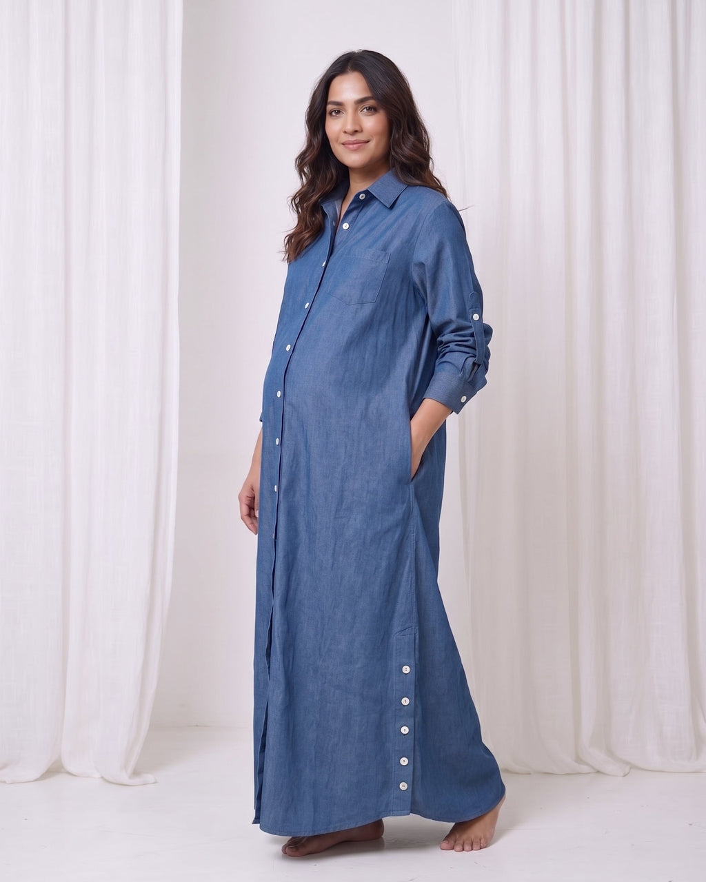 A woman modeling a long-sleeved, button-front maxi shirt dress in a dusty blue denim-like fabric, standing against a white draped curtain backdrop.