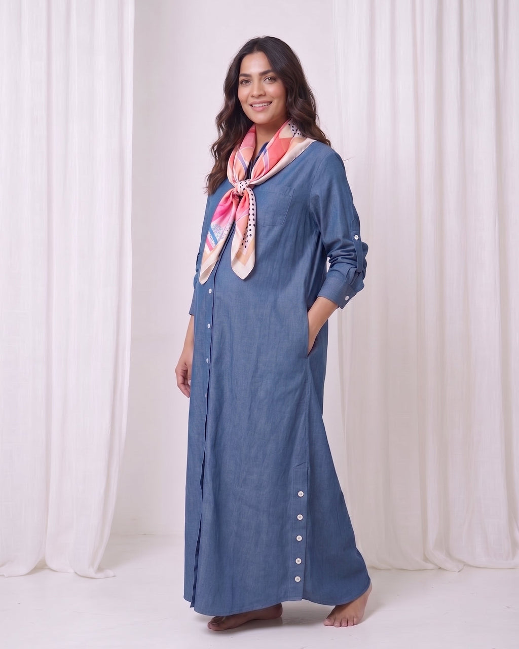 A woman modeling a long-sleeved, button-front blue maxi dress with side pockets, styled with a pink and white patterned scarf, standing against a white draped curtain backdrop.