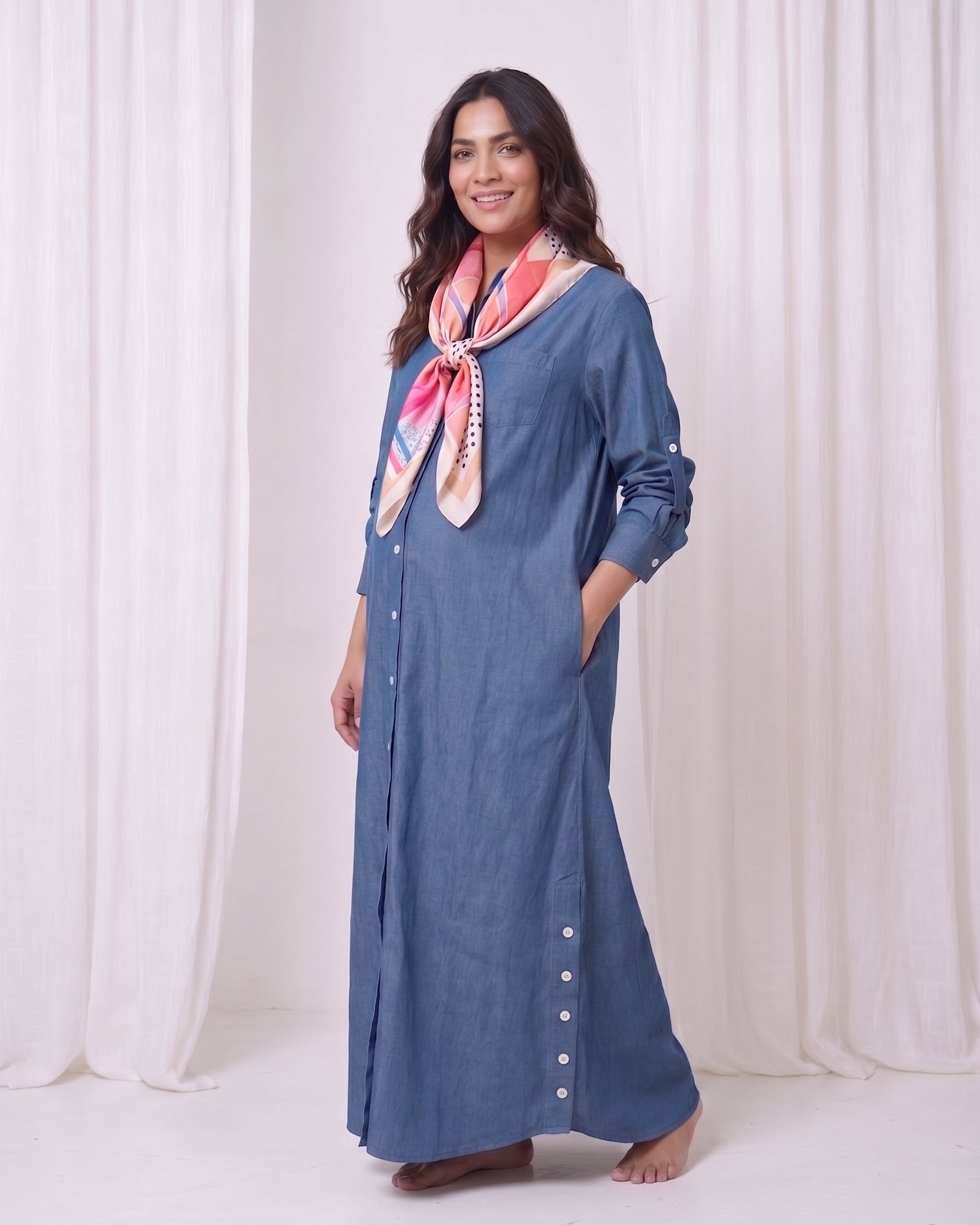 A woman modeling a long-sleeved, button-front blue maxi dress with side pockets, styled with a pink and white patterned scarf, standing against a white draped curtain backdrop.